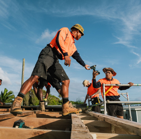 Skilled military tradespeople work on the roof of a building. One soldier hands a drill to another under blue skies,