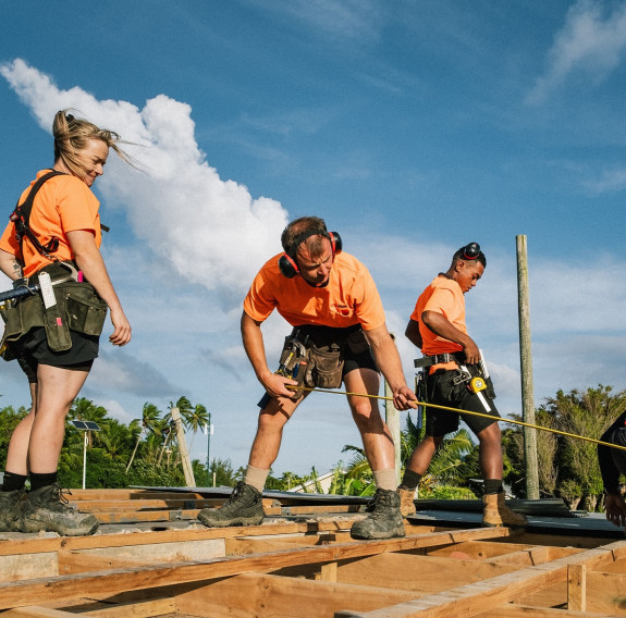 Four skilled military tradespeople work in a roof in front of puffy white clouds and blue skies. One soldier extends a tape measure across a beam.