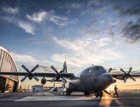 C-130H(NZ) Hercules stationary on flightline at RNZAF Base Auckland. With hangars in the background, a large puddle mirrors the aircraft and blue sky above.